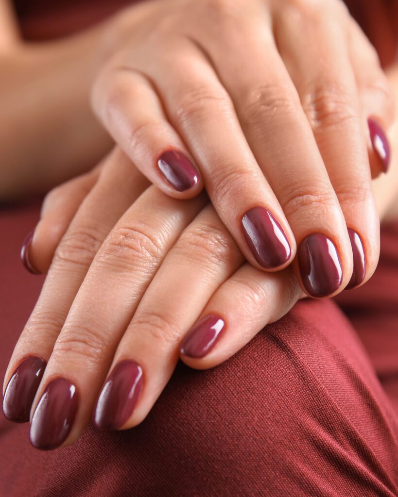 Two hands with short round nails in glossy plum-burgundy gel polish resting on rust-red fabric