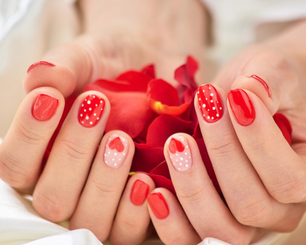 Two hands with coral nails decorated with white dots and hearts, resting on a red petal
