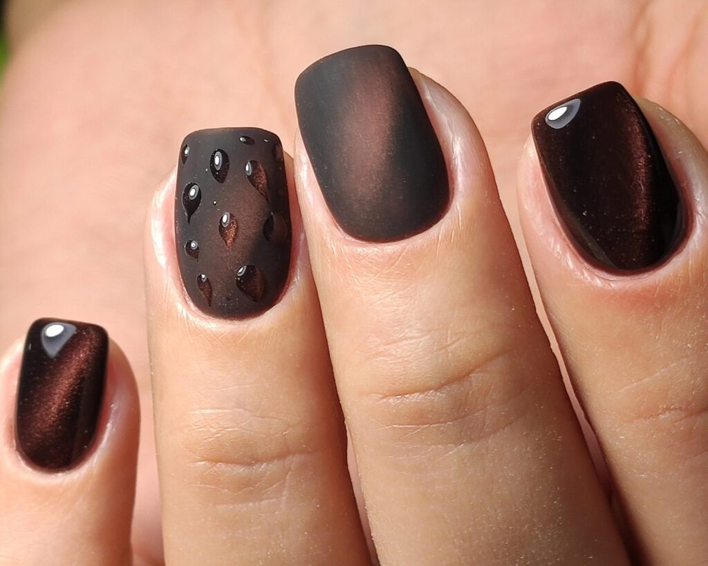 Close view of one hand with nails in matte and glossy dark brown, one set with small stones