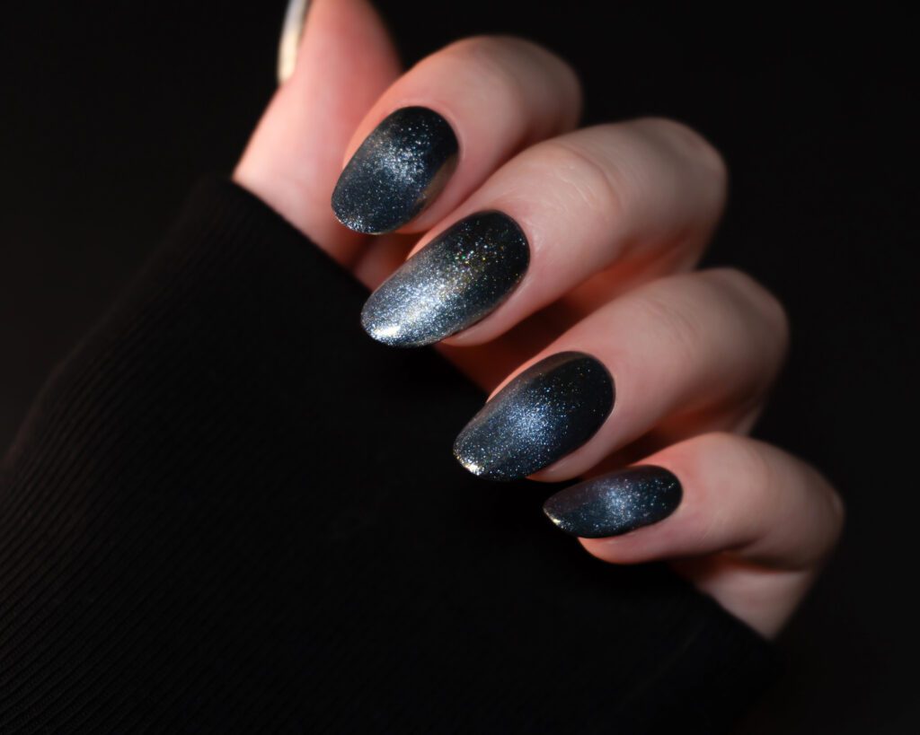 A hand against black with almond nails in a dark teal glitter gradient