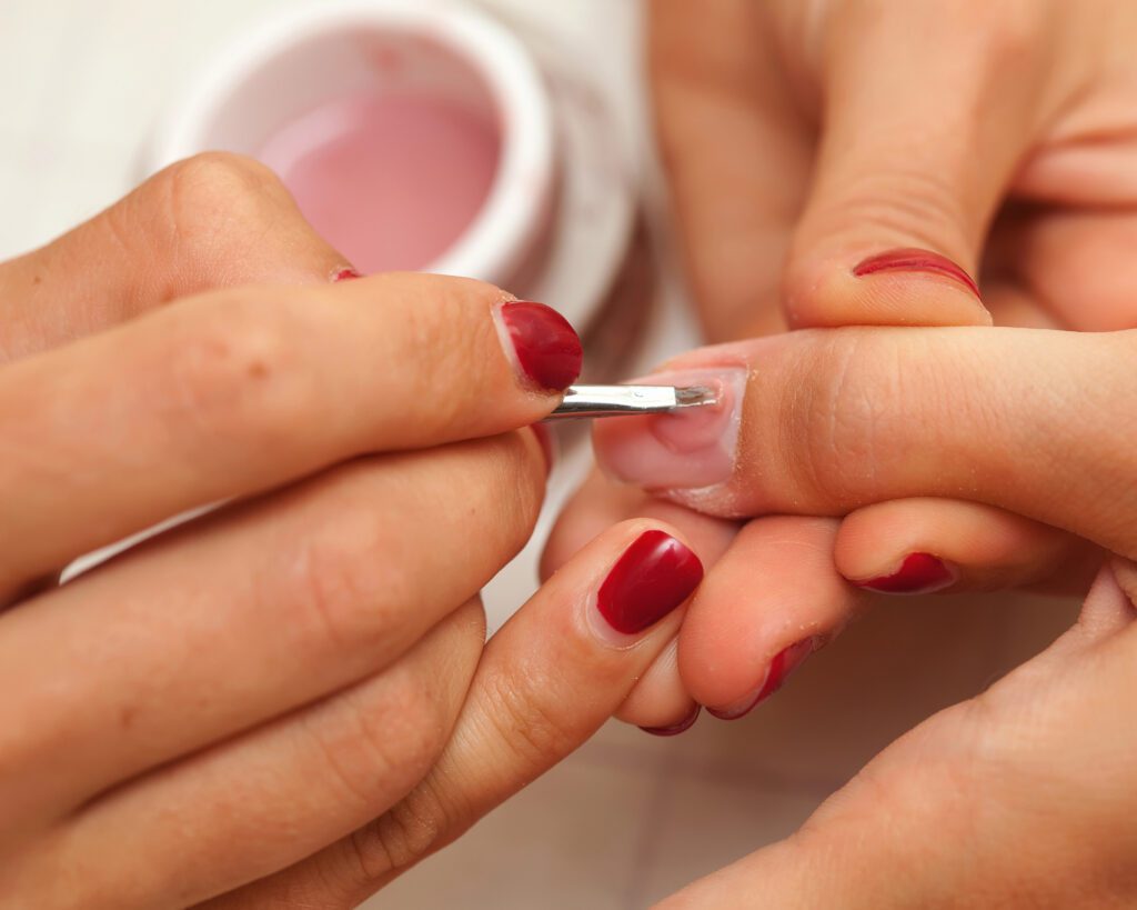 Technician brushing clear builder gel onto a natural nail beside a pot of pink gel