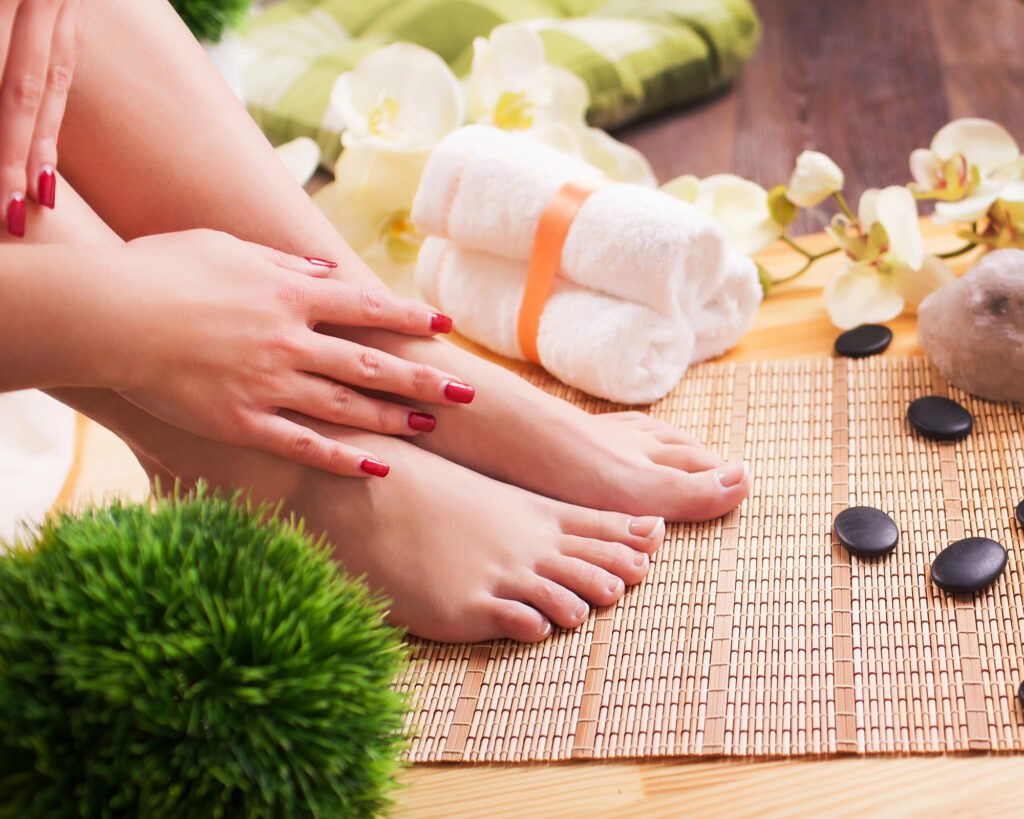 Bare feet and hands with red nails on a bamboo mat, with rolled towels, orchids and smooth stones