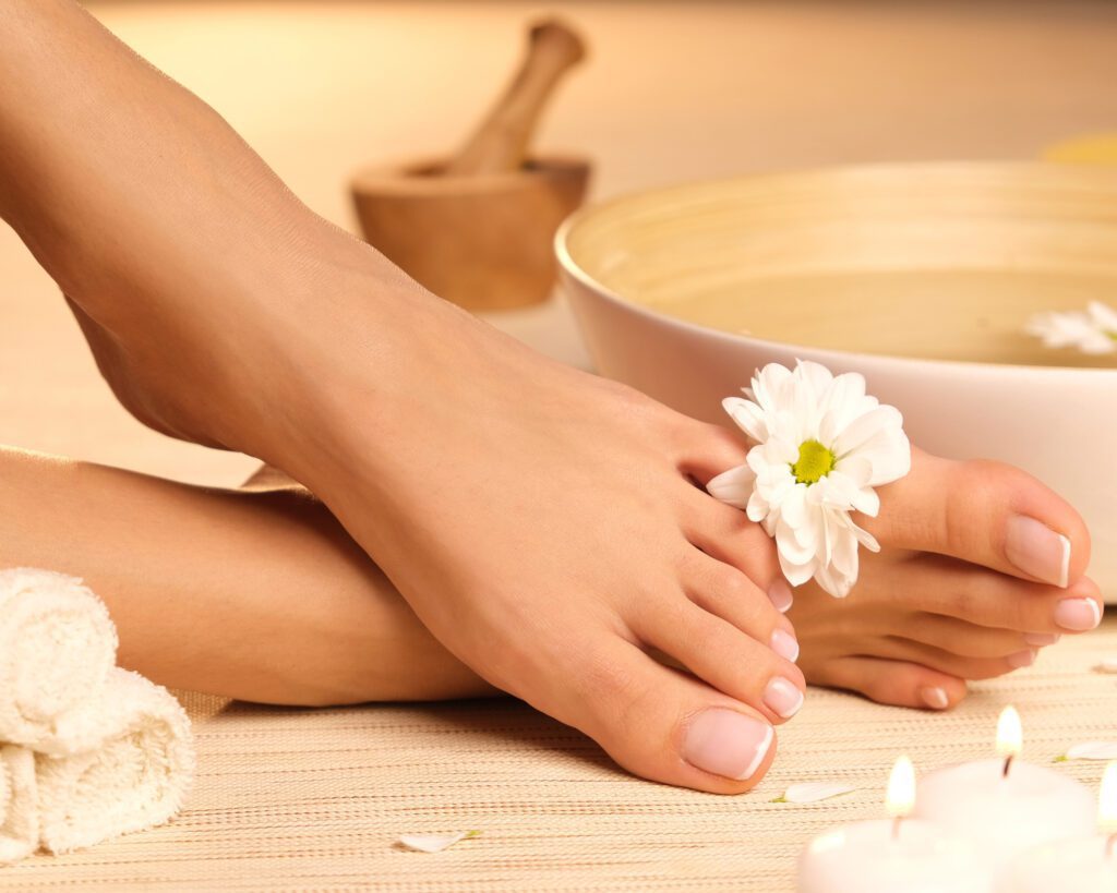 A foot with natural toenails resting on a wooden surface beside a white daisy, a bowl and a wooden scoop