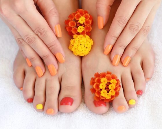 Hands and feet with alternating red, orange and yellow nails beside orange flowers