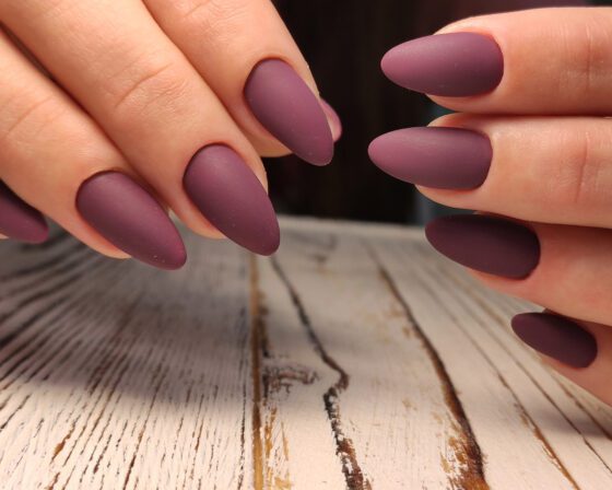 Close view of fingers from two hands above pale wood, showing almond nails in a muted matte plum