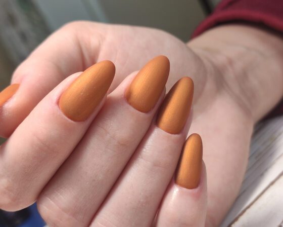 Close-up of a hand against pale textured and burgundy fabric, showing medium matte almond nails in metallic copper