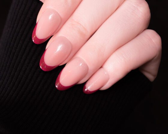 Tight close-up of one hand against dark ribbed fabric, showing medium blush almond nails with deep red French tips