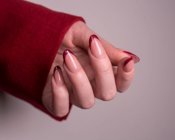 Vertical close-up of curled hand with a red sleeve against a muted gray background, showing medium blush almond nails with deep red French tips