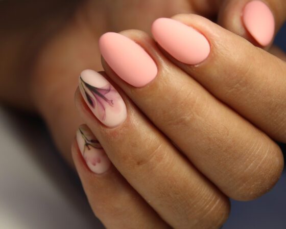 Extreme close-up of a curled hand against a warm blurred background, showing matte peach almond nails with fine dark botanical sketches on sheer accent nails