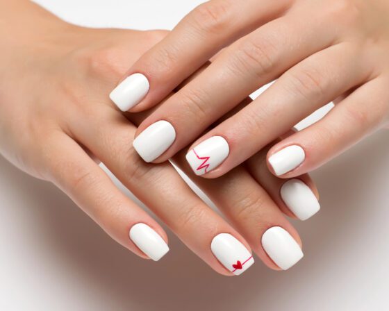 Close-up of overlapping hands on a bright pale background, showing short white nails with a fine red heartbeat line and a small red heart on accent nails