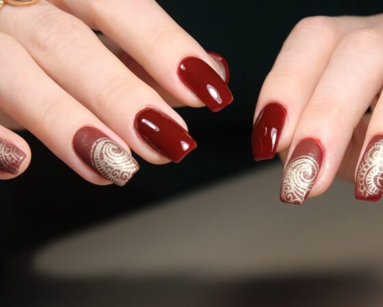Close view of two hands against a dark background, showing glossy burgundy nails with cream and gold scrollwork on accent nails