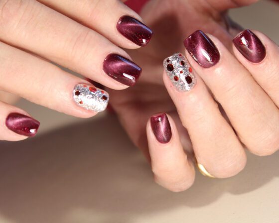 Close-up of two hands against a pale neutral background, showing wine red nails with a luminous sheen and a silver glitter accent set with small red and gold stones