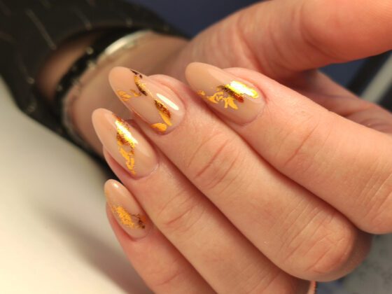 Macro close-up of overlapping hands on a pale surface, showing medium sheer nude almond nails with irregular metallic gold flakes