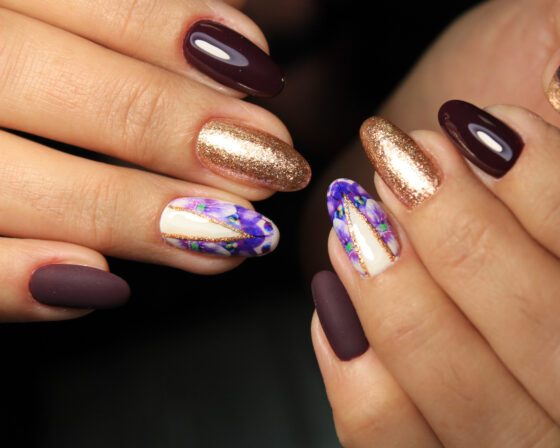 Extreme close-up of two hands against a dark background, showing almond nails mixing glossy and matte plum, copper glitter and violet-marbled white accents
