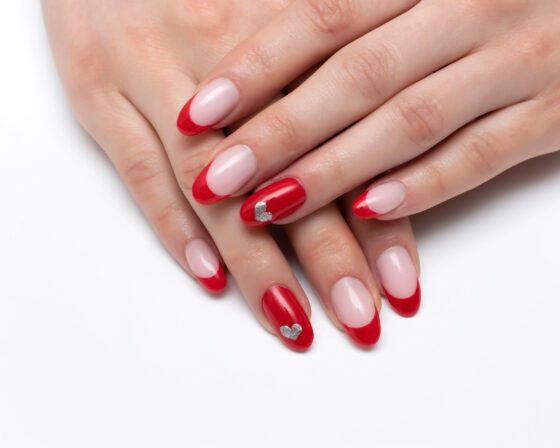 Close-up of crossed hands on white, showing pale pink nails with bright red French tips and solid red accent nails with small silver details