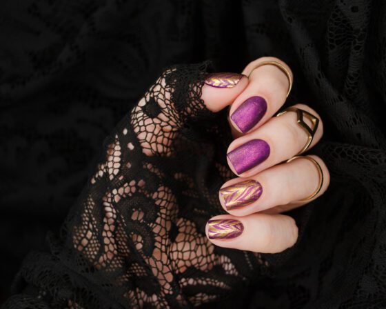 Vertical close-up of a hand curled against black lace, showing short violet shimmer nails with gold chevron patterns on accent nails