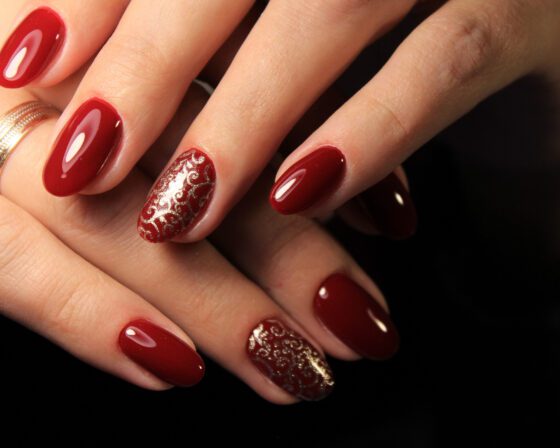 Close crop of overlapping hands against a nearly black background, showing glossy deep red nails with gold filigree on one accent nail per hand