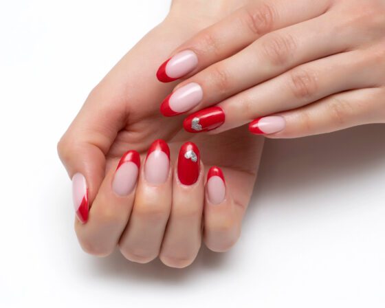 Close view of hands in a compact folded pose against white, showing pale pink nails with bright red French tips and solid red accent nails with small silver details