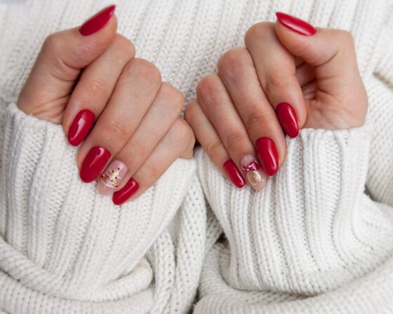Two curled hands resting against a white knit sweater, showing glossy red almond nails with a pale accent carrying a small gold detail