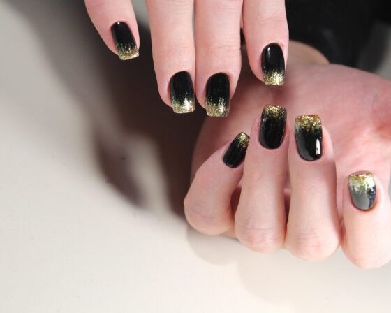 Two hands against a pale neutral background, showing glossy black nails with gold glitter fading in from the tips