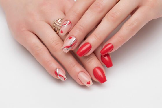 Close-up of overlapping hands on a pale neutral background, showing bright red nails with nude accent nails carrying fine silver curves and tiny red hearts