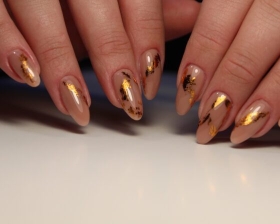 Two hands arranged above a pale reflective surface, showing medium sheer nude almond nails with irregular metallic gold flakes
