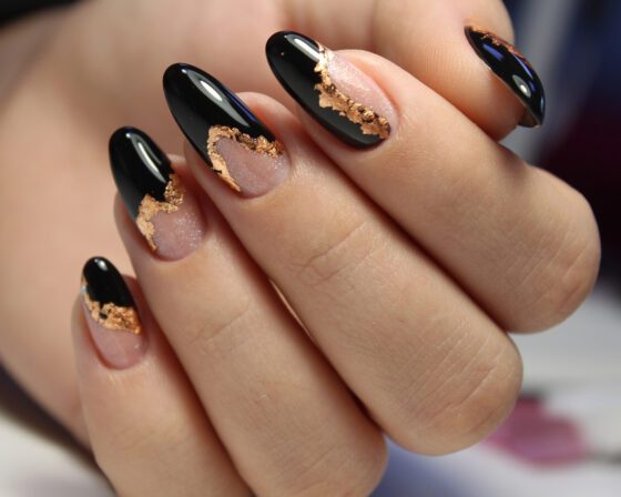 Macro close-up of curled fingers against a dark backdrop, showing glossy black and sheer nude almond nails edged with metallic gold flakes