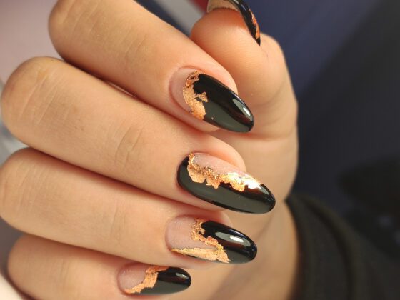 Macro close-up against a dark blurred background, showing glossy black and sheer nude almond nails edged with metallic gold flakes