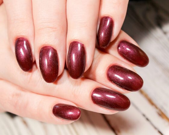Close crop of overlapping hands over a pale wood surface, showing medium oval nails in a deep wine red with a fine metallic shimmer
