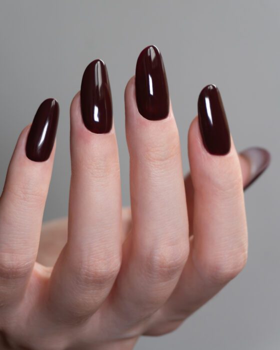 Vertical close-up of raised fingertips against a gray seamless background, showing medium almond nails in a glossy deep burgundy shade