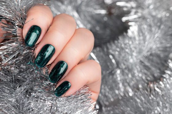 Close crop of a curled hand nestled into silver tinsel, showing short nails in a deep teal green with fine sparkle