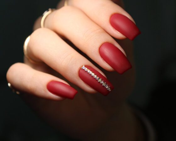 Close portrait of a curled hand against a dark background, showing medium matte berry-red nails with a slim line of crystals on one accent nail