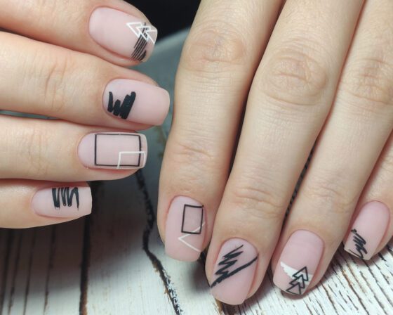 Close-up of two hands on pale wood, showing short matte blush nails with black brush marks, outlined squares and fine white lines