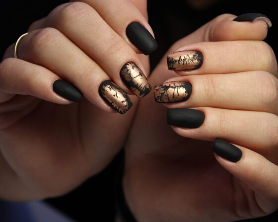Two hands close together against a dark backdrop, showing matte black nails alternating with distressed metallic gold accents