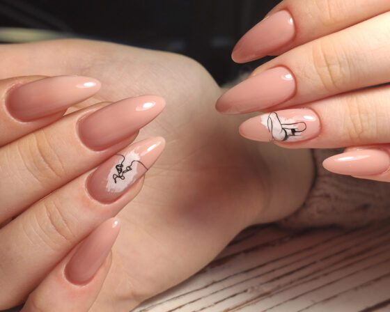 Extreme close-up of overlapping fingers above pale wood, showing long glossy peach-nude almond nails with small black line drawings on accent nails
