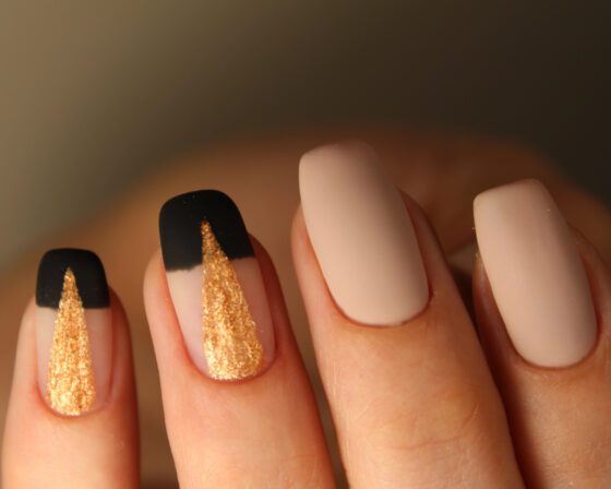 Tight macro of raised fingertips against a warm dark background, showing medium matte nude nails with black tips and gold glitter panels on accent nails