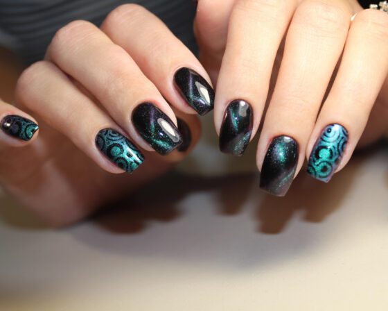 Two hands close together against a pale neutral background, showing dark navy-violet shimmer nails with turquoise circle patterns on accent nails