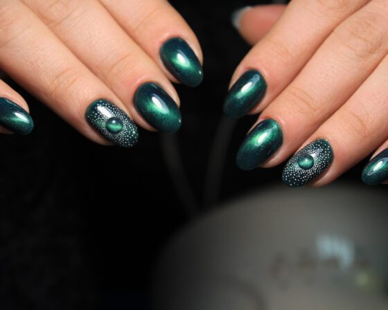 Close horizontal crop of hands against a black background, showing deep teal almond nails with a reflective sheen and a dotted green-stone accent