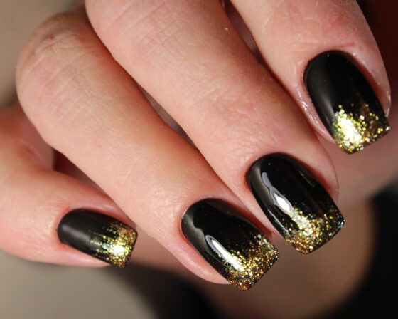 Tight macro crop of fingers against a neutral background, showing glossy black nails with gold glitter fading in from the tips
