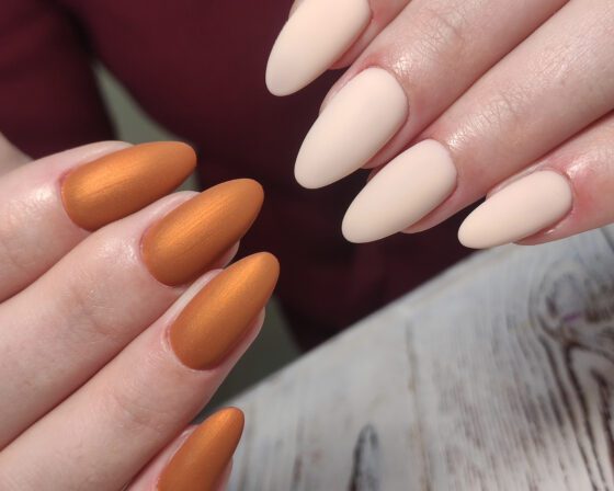 Close-up of two hands over pale wood and burgundy fabric, showing medium matte almond nails alternating metallic copper and cream