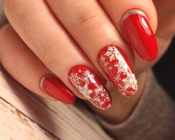 Tight close-up of fingers against pale wood and dark fabric, showing medium glossy red nails with white snowflake patterns on accents and silver sparkle near the base