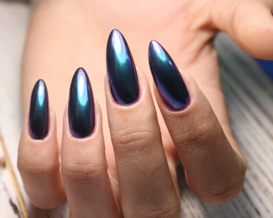 Extreme close-up of one hand over a pale wood surface, showing long pointed navy nails with a turquoise and violet colour-shifting sheen