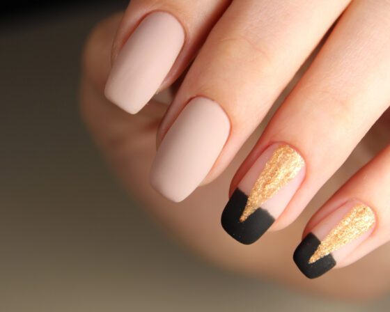 Macro crop of fingertips against a dark warm background, showing medium matte nude nails with black tips and gold glitter panels on accent nails