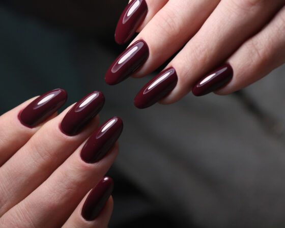 Tight view of crossed hands against a dark blurred background, showing medium almond nails in a glossy deep burgundy shade