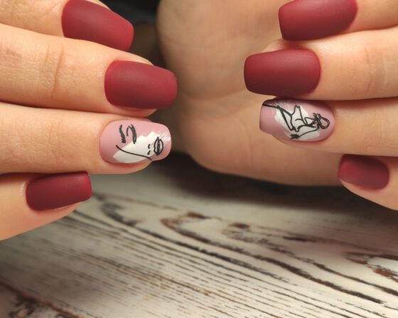 Horizontal close-up of facing hands above pale wood, showing matte berry-red nails with pale pink accents carrying black line drawings
