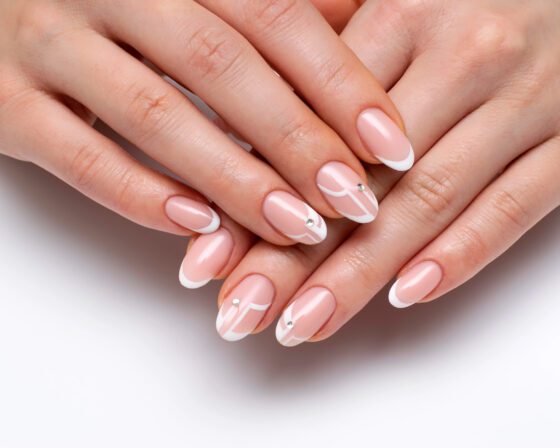 Close-up of crossed hands on a bright white background, showing pale pink nails with white French tips, fine white line art and a tiny crystal