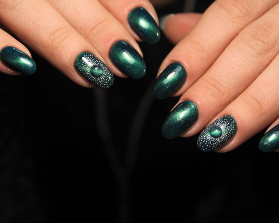 Tight view of two hands against a black background, showing deep teal almond nails with a reflective sheen and a dotted green-stone accent
