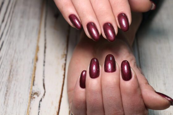 Two hands arranged above a pale wood surface, showing medium oval nails in a deep wine red with a fine metallic shimmer