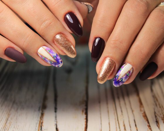 Close horizontal crop of two hands above a pale wood surface, showing almond nails mixing glossy and matte plum, copper glitter and violet-marbled white accents