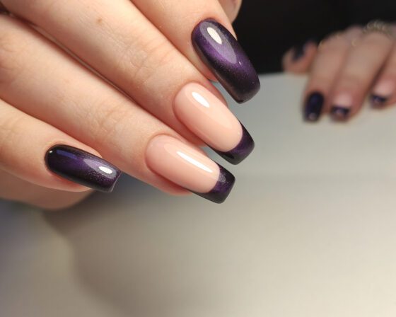 Macro view of overlapping hands against a neutral gray background, showing long nude nails with deep violet shimmer French tips